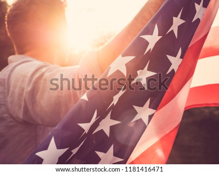 Attractive man holding waving in the wind US flag against the backdrop of the rays of the setting sun. View from the back, closeup. Preparing for the holidays