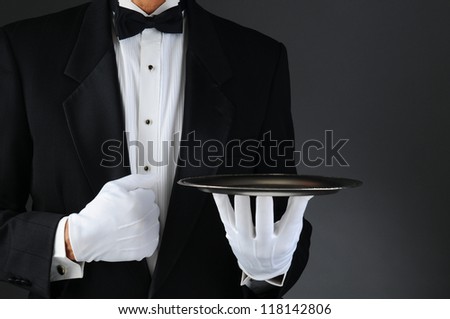 Closeup of a tuxedo wearing waiter holding a silver tray in front of his body. Horizontal format on a light to dark gray background. Man is unrecognizable. Royalty-Free Stock Photo #118142806
