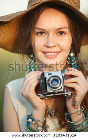 Happy woman holding an old camera in her hand. A beautiful smiling hipster girl with bracelets decorating her arms in a wide-brimmed straw hat uses an old camera. Close-up.
