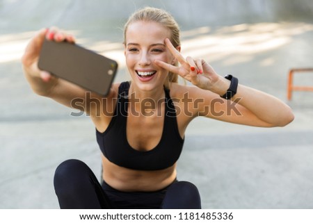 Image of amazing young sports woman sitting outdoors take a selfie by mobile phone showing peace gesture.