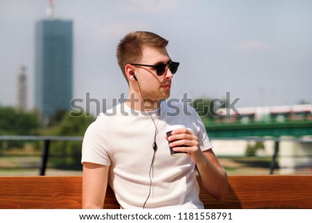 Handsome boy wearing sunglasses using tablet and headphones, sitting on a bench and drinking coffee to go. Handsome student immersed in social media. Background scenery bridge, river and green grass.