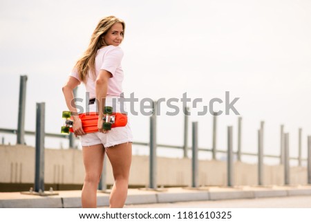 Portrait of Young Beautiful Smiling Blonde Girl with Orange Skateboard on the Bridge