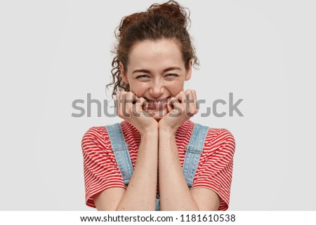 Funny freckled teenager laughs at good joke, keeps hands under chin, grins from laughter, has freckled skin, curly dark hair, dressed in casual t shirt and overalls, stands against white background