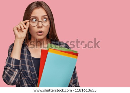 Surprised teenager looks with shocked expression aside, dressed in casual outfit, uses literature for preparing to exams, isolated over pink wall with copy space for your advertising content