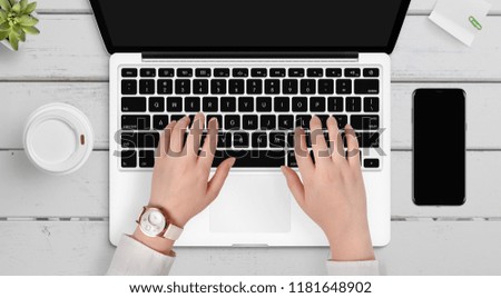 Female hands working on modern laptop at the white wooden desk surrounded with phone, coffee, plant and note paper