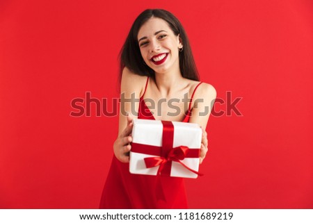 Portrait of a smiling young woman in dress holding present box isolated over red background