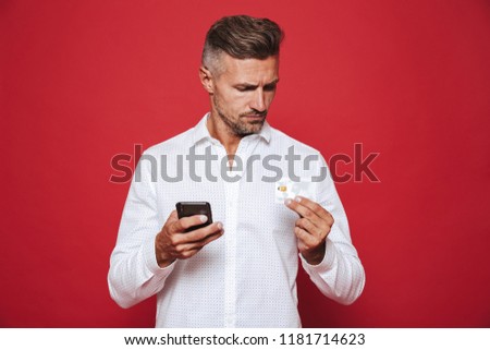 Photo of european man in white shirt holding credit card and smartphone isolated over red background
