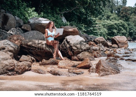 Young american woman in blue swimsuit working with laptop on empty beach and sitting on stone. Concept of resting on morning sea and summer vacations, modern technology.