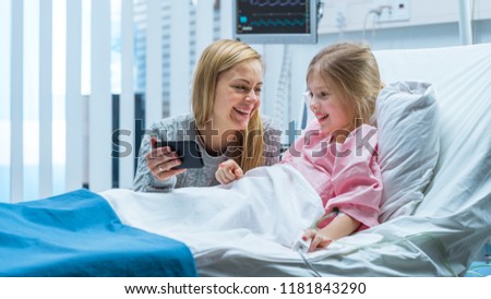 Cute Little Girl Lies on a Bed in the Children's Hospital, Her Mother Sits Beside, They Watch Cartoons/ Funny Videos on Smartphone. Modern Pediatric Ward. Royalty-Free Stock Photo #1181843290