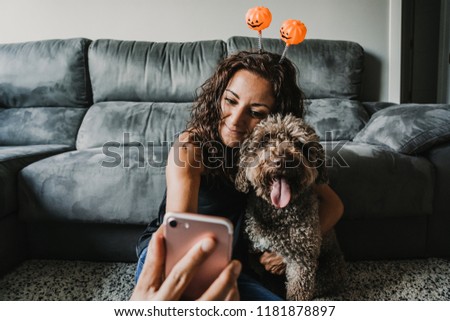 
Young and cheerful woman sitting on the sofa of her house taking some pictures with her smartphone and her funny dog. With Halloween costumes. Lifestyle.