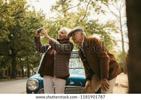 Senior couple on road trip taking pictures of nature landscape with digital camera. Old woman taking photographs with her camera with man standing by and looking at camera.