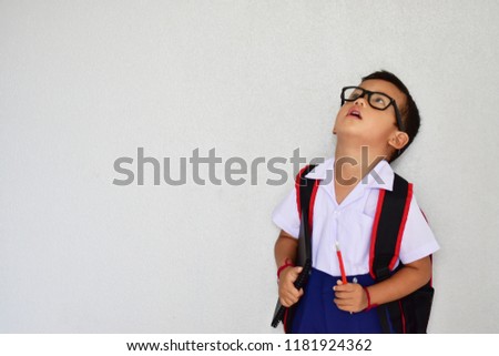 Back to school, little nerdy Asian kindergarten boy in school uniform wearing glasses carrying a bag and a notebook ready to go to school on light grey background, first day to school