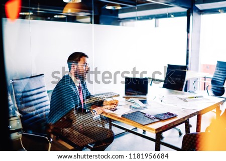 Professional male banker sitting at his loft interior cabinet with glass walls working on business planning, confident owner in formal suit signing contracts in office sitting at desktop with netbook