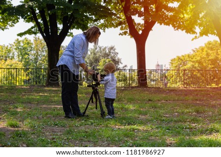 Two years old boy is photographer. Kid holds a camera on tripod and takes photo of landscape. Little child dressed shirt and jeans playing in the park.