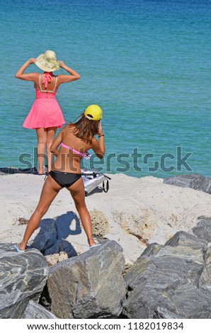 Young woman wearing a black and pink bikini taking a cell phone photo of aa woman wearing a pick beach dress and a straw sun hat