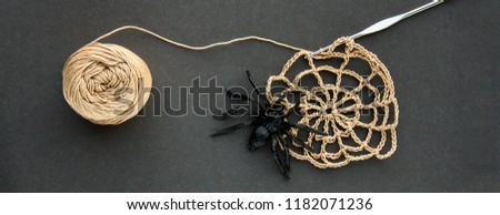 Crocheted cobweb with black rubber spider, crochet and cotton yarn ball of threads on black paper background. Dark halloween october concept. Top view. Web banner