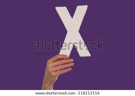 Female hand holding up the uppercase capital letter x isolated against a purple background conceptual of the alphabet, writing, literature and typeface