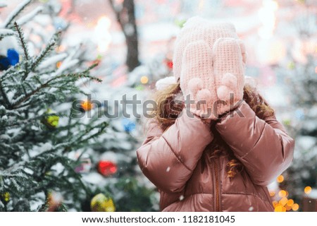christmas portrait of happy kid girl playing outdoor in snowy winter day, fir trees decorated for New Year holidays with toys and lights on background