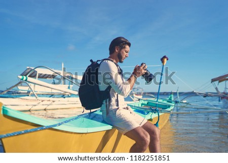 Photography and travel. Young man with rucksack taking photo with his camera enjoying beautiful tropical sea view on fishing beach.