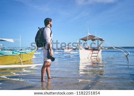 Photography and travel. Young man with rucksack holding camera walking on fishing beach enjoying beautiful tropical sea view.