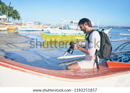 Photography and travel. Young man with rucksack taking photo with his camera enjoying beautiful tropical sea view on fishing beach.