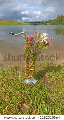 Golden alto saxophone on a background of nature. Original saxophone with a bouquet of summer summer flowers. Art. Cover for jazz. Reflection in water. unexpected concept