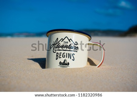 Mug saying "adventure begins" with a logo with a person canoeing placed in sand