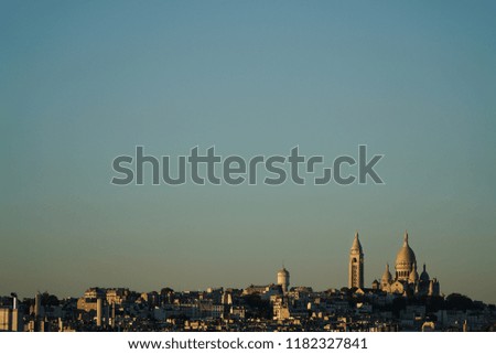 Minimal landscape view over Montmartre and Paris skyline, with several roofs and buildings in first plan and Sacre Coeur church in background, under the warm golden light of summer sunset, France.
