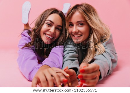 Beautiful european sisters lying on the floor and smiling. Indoor photo of good-humoured female friends chilling together.