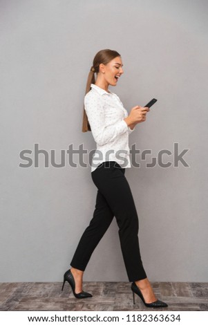 Image of beautiful smiling business woman walking over grey wall background using mobile phone.