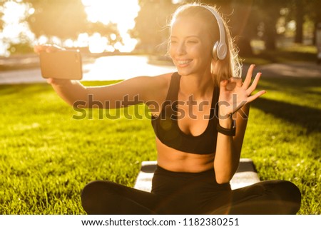 Photo of beautiful young blonde sports woman in park outdoors listening music with headphones take a selfie by mobile phone waving.
