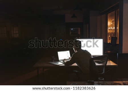 Rearview of a young businessman sitting alone at a desk in a dark office working on a laptop late in the evening Royalty-Free Stock Photo #1182383626