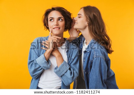 Photo of cute young women friends posing isolated over yellow background gossiping.