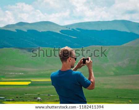 Young blonde man in sunglasses takes pictures on his mobile phone Landscape of green hills and endless fields divided into squares in Castelluccio di Norcia