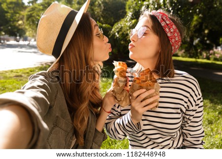 Two lovely young girls friends having fun at the park, taking a selfie, holding croissants