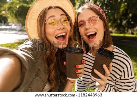 Two excited young girls friends having fun at the park, taking a selfie, holding coffee cups