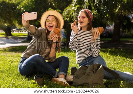 Two joyful young girls friends having fun at the park, taking a selfie, having a picnic