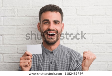 Young adult man over brick wall holding blank card screaming proud and celebrating victory and success very excited, cheering emotion