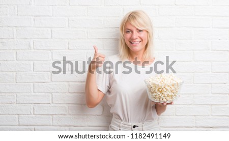 Adult caucasian woman over white brick wall eating pop corn happy with big smile doing ok sign, thumb up with fingers, excellent sign