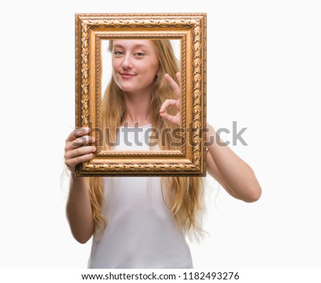 Blonde teenager woman holding vintage frame art doing ok sign with fingers, excellent symbol