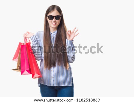 Young Chinese woman over isolated background holding shopping bags on sales doing ok sign with fingers, excellent symbol