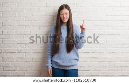 Young Chinise woman over white brick wall showing and pointing up with finger number one while smiling confident and happy.
