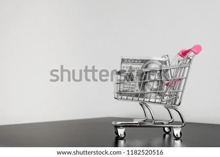 Pink and metal shopping cart on black table on white background with  one hundred dollars banknote.  Minimalism style. Shop trolley at supermarket. Sale, discount, shopaholism concept