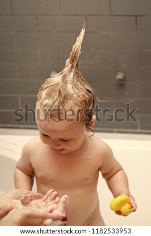 Small pretty male kid standing in bathroom with wet foam hair holding yellow duckling toy looking at hands of mother, vertical picture
