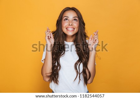 Portrait of a smiling young girl with long brunette hair standing over yellow background, holding fingers crossed for good luck Royalty-Free Stock Photo #1182544057
