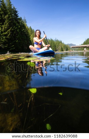 Over and Under picture of a woman paddle boarding during a vibrant sunny summer day. Taken in Cedar Lake, Northern Vancouver Island, British Columbia, Canada.