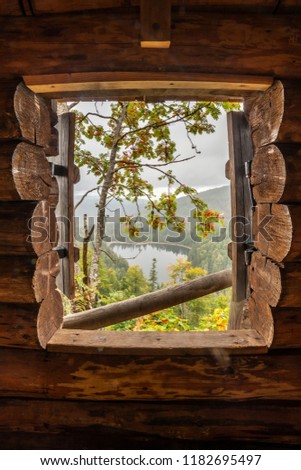 View through the window of chapel on a ledge into the foggy valley with a lake in the middle of endless forest. The horizon is covered in clouds and fog and a mountain ash shows its red berries.