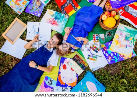 Authentic artist children in blue aprons girl and boy paints lying in creative chaos palette watercolor paints palette and brush . Painting outdoor grass autumn.Top view handmade crafts.