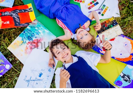 Authentic artist children in blue aprons girl and boy paints tickle each other by paint brushes, lying in creative chaos palette watercolor paints, brush, drawings.Painting outdoor. Top view