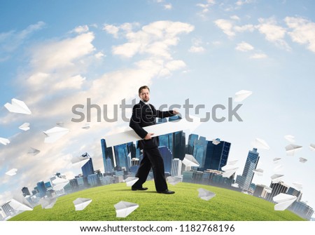 Confident and young businessman in suit holding big white arrow in hands which pointing to the side while standing among flying paper planes on green lawn and cityscape view on background.